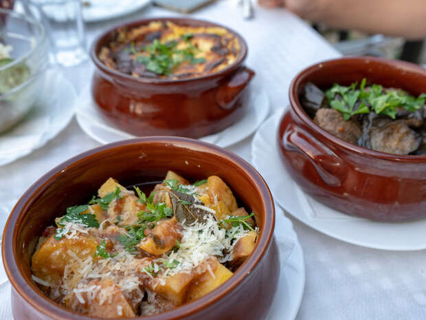 Clay bowls contain various cooked dishes topped with herbs and cheese, resting on a white tablecloth, suggesting an outdoor dining setting with partially visible plates, glassware, and a person’s arm nearby.
